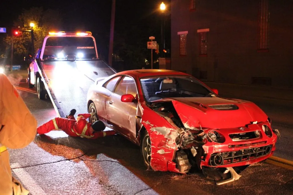 A red car with severe front-end damage, being pulled onto a tow bed.