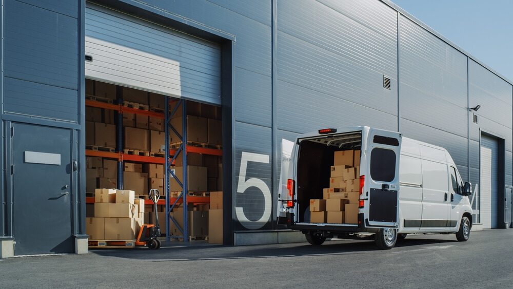 Outside of Logistics Warehouse with Open Door, Delivery Van Loaded with Cardboard Boxes