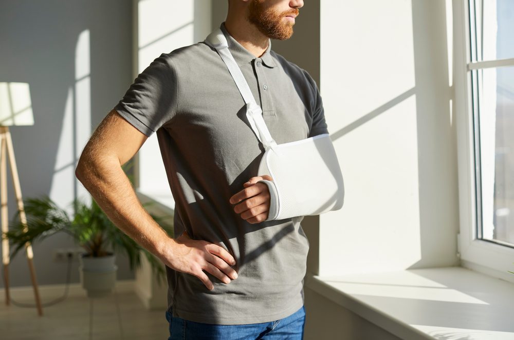 A man wearing a gray polo, with his hand in a sling, looking out a window.